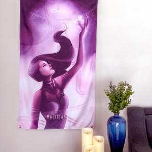 May include: A purple wall tapestry featuring a woman with long dark hair reaching towards a glowing orb. The text "THE MAGICIAN" is at the bottom. The tapestry is hanging on a white wall, next to a blue vase with greenery and three ivory candles.