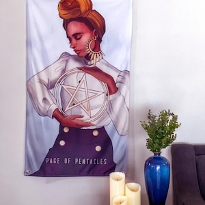 Puede incluir: Un tapiz de pared con una ilustración de una persona sosteniendo un pentáculo. La persona lleva una blusa blanca, una falda morada oscura y un turbante. El texto "PAGE OF PENTACLES" está impreso en la parte inferior. El tapiz está colgado en una pared blanca.