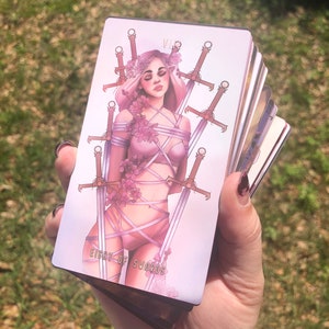 Puede incluir: Una baraja de cartas del tarot con la carta de los Ocho de Espadas. La carta representa a una mujer atada por espadas y detalles florales, con una paleta de colores rosa suave. La carta se sostiene en una mano sobre un fondo verde borroso.