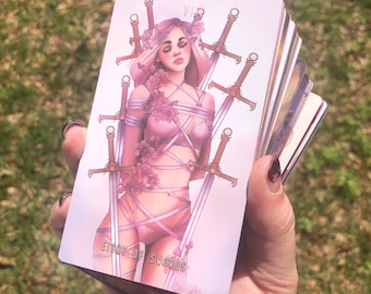 May include: A tarot card deck featuring the Eight of Swords. The card shows a woman bound by swords and floral accents, in a soft pink colour scheme. The card is held in a hand against a blurred green background.