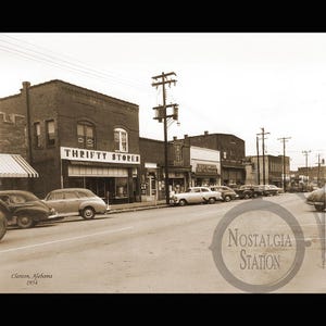 May include: Black and white vintage photograph of a street scene in Clanton, Alabama, from 1954. The image shows classic cars parked along the street in front of brick buildings, including a store with the sign "Thrifty Stores".