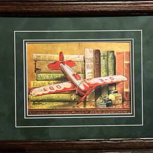 May include: A vintage toy airplane with red and silver paint is resting on a stack of books. The books have titles like "Aircraft History", "Flight School", "Flying High", and "Sky Racers". The airplane has a string attached to it and the number "G-E 087" is visible on the side.