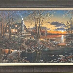 May include: A framed landscape painting depicts a cabin nestled among trees on a lake at sunset. The cabin has a stone chimney with smoke, and a wooden dock extends into the water. The sky is a mix of orange, yellow, and blue.
