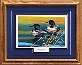 May include: Framed print of two loons with a chick on a lake. The loons have black heads and white-spotted backs. The water reflects the birds and the colorful sky. The frame is wood with a blue mat and the print is titled "Calm Reflections".