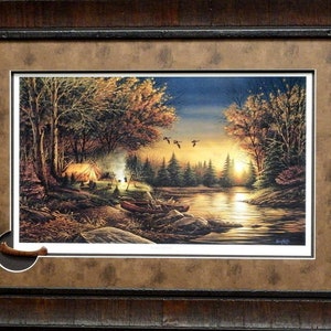 May include: A framed print of a painting depicting a campsite in a forest by a lake at sunset. The scene features a tent, a campfire, and a canoe. There are trees and a lake in the background. The sky is a deep blue with orange and pink hues. The print is matted and framed in a dark wood frame.