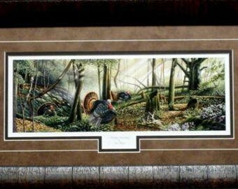 Op de afbeelding: Een ingelijste print van een schilderij dat een bos scène toont met wilde kalkoenen in het bos. De kalkoenen zijn bruin en zwart met rode koppen en lopen door de bomen. De achtergrond is een weelderig groen bos met zonlicht dat door de bomen schijnt.