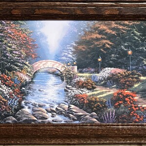 May include: A framed landscape print featuring a serene garden scene. A stone bridge arches over a flowing stream, surrounded by lush greenery, colorful flowers, and trees. The artwork is enclosed in a dark brown wooden frame.