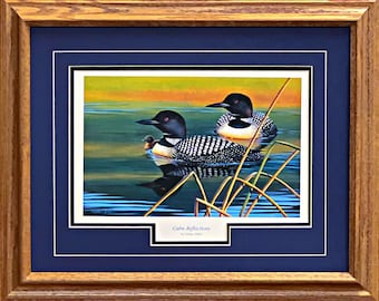 May include: Framed print of two loons with a baby loon swimming on a lake. The loons have black heads and white-spotted black bodies. The water reflects the birds and the colorful sky. The print is in a blue mat and a wooden frame. The text "Calm Reflections" is at the bottom.