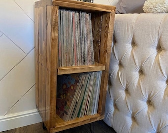 May include: A wooden record stand with two shelves, filled with vinyl records. The stand has hairpin legs and is stained a dark brown color.