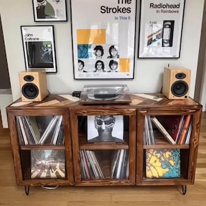 May include: Wooden record cabinet with glass doors, holding vinyl records. A turntable sits on top, flanked by two wooden speakers. The cabinet has a geometric design on top and is set against a wall with framed band posters.