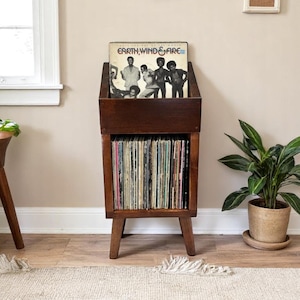 May include: A dark brown wooden record player stand with a shelf for storing records. The stand has a top shelf with a record on it, featuring the album cover for "Earth, Wind & Fire".