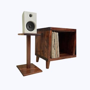 May include: A white speaker on a dark wood speaker stand next to a dark wood record player stand with a shelf of vinyl records.