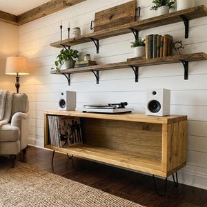 May include: A rectangular wooden media console with black metal hairpin legs, featuring an open shelf containing vinyl records. On top sits a white turntable flanked by two white speakers. The console is positioned against a white shiplap wall.