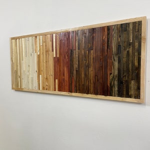 May include: A rectangular wall art piece made of reclaimed wood with a natural wood frame. The wood is arranged in horizontal strips of varying widths and colors, creating a gradient effect from light to dark. The colors range from light beige to dark brown, with shades of red and orange in between.