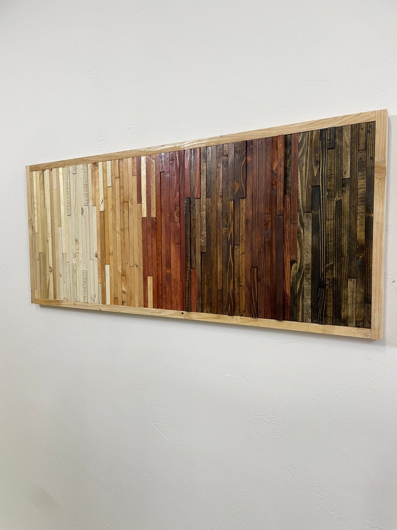May include: A rectangular wall art piece made of reclaimed wood with a natural wood frame. The wood is arranged in horizontal strips of varying widths and colors, creating a gradient effect from light to dark. The colors range from light beige to dark brown, with shades of red and orange in between.