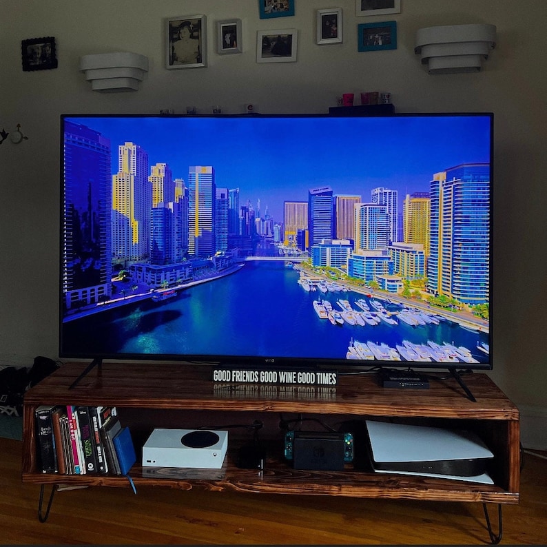 May include: A dark wood entertainment center with a flat screen TV displaying a cityscape with a body of water in the foreground. The TV is on a shelf with a white gaming console, a black gaming controller, and a white rectangular box. The text "GOOD FRIENDS GOOD WINE GOOD TIMES" is displayed on the shelf below the TV.