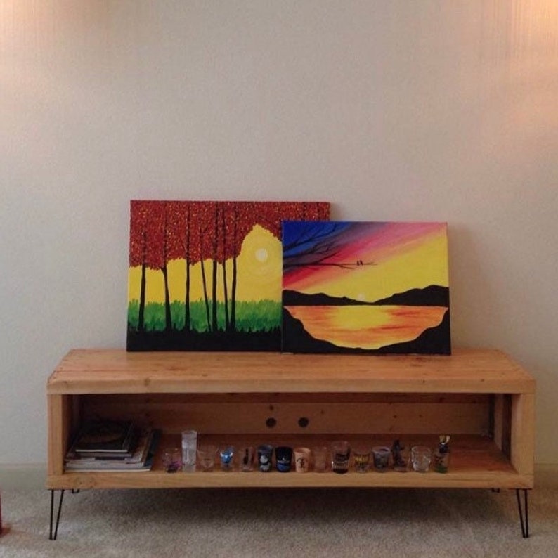 May include: A wooden shelf unit with two paintings on top. The paintings are both landscapes, one with a sunset and the other with trees. The shelf has a lower shelf with shot glasses and books.