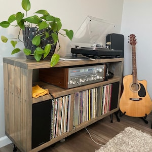May include: A wooden record player stand with a record player, amplifier, and speakers. The stand has a shelf for records and a plant on top. There is a guitar leaning against the stand.