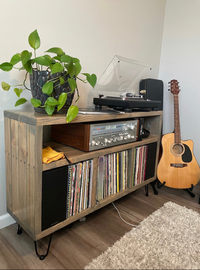 May include: A wooden record player stand with a record player, amplifier, and speakers. The stand has a shelf for records and a plant on top. There is a guitar leaning against the stand.
