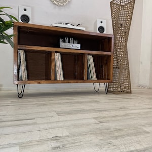 May include: A wooden record player stand with three shelves for vinyl records. The stand has black hairpin legs and is sitting on a light wood floor. There are two white speakers on the top shelf, a record player, and an amplifier.