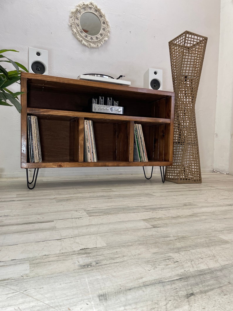 May include: A wooden record player stand with three shelves for vinyl records. The stand has black hairpin legs and is sitting on a light wood floor. There are two white speakers on the top shelf, a record player, and an amplifier.