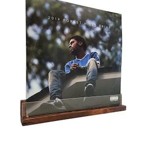 May include: A brown wooden record stand with a vinyl record of the album "2014 Forest Hills Drive" by J. Cole. The album cover features a man sitting on a rooftop with a blurred background of trees and sky.
