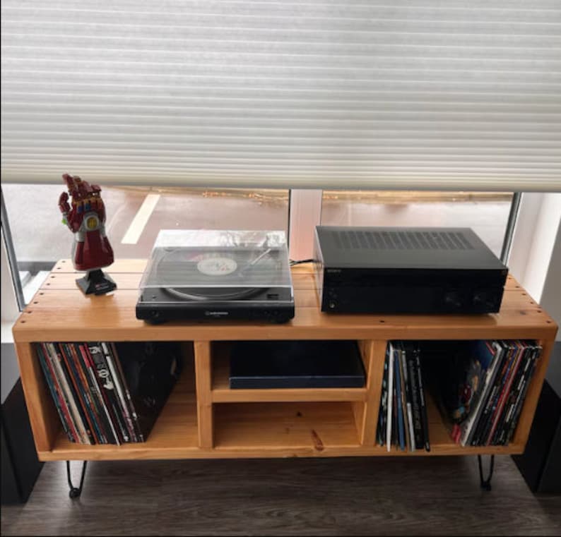 May include: A wooden record player stand with three shelves. The top shelf has a black record player with a clear lid, a black amplifier, and a red Iron Man hand figurine. The bottom shelves are filled with vinyl records.