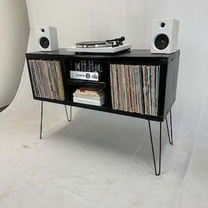 May include: Black record player stand with two white speakers, a turntable, and shelves for vinyl records. The stand has black hairpin legs.