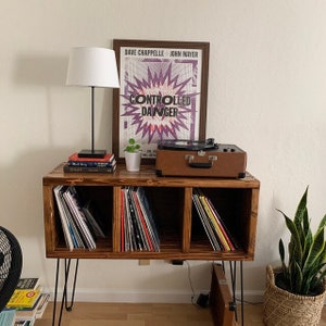 K&ouml;nnte beinhalten: Ein Holz-Schallplattenst&auml;nder mit drei Regalen f&uuml;r Schallplatten. Der St&auml;nder hat schwarze Haarnadelbeine und ist mit einem Vintage-Schallplattenspieler, einer Lampe und einem Poster mit der Aufschrift "Dave Chappelle John Mayer Controlled Danger" dekoriert.