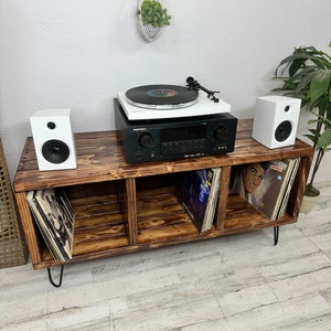 May include: A dark brown wooden record player stand with three shelves. The stand has hairpin legs and is holding a turntable, a stereo receiver, and two white speakers. There are vinyl records on the shelves.