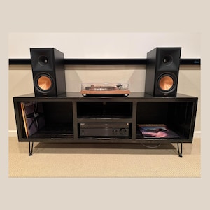 May include: A black entertainment center with a turntable, amplifier, and two black speakers with copper-colored cones. Vinyl records are visible in the open compartments. The unit has black hairpin legs and is set against a neutral background.