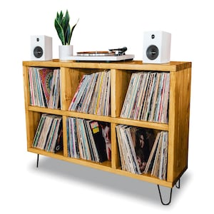 May include: A wooden record shelf with six compartments, each filled with vinyl records. The shelf has a light wood finish and sits on hairpin legs. Two white speakers are placed on top of the shelf, and a turntable is in the center.