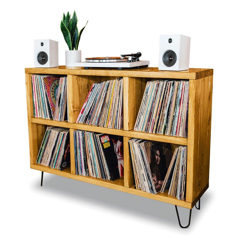 May include: A wooden record shelf with six compartments, each filled with vinyl records. The shelf has a light wood finish and sits on hairpin legs. Two white speakers are placed on top of the shelf, and a turntable is in the center.