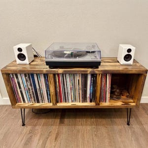 May include: A wooden record player stand with three shelves for storing vinyl records. The stand has hairpin legs and is painted a dark brown color. A turntable sits on the top shelf, and two white speakers are on either side of the turntable.