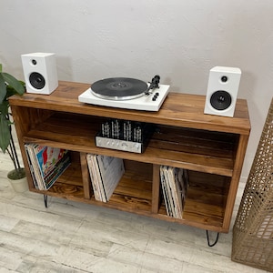 May include: A dark brown wooden record player stand with three shelves for storing vinyl records. The stand has black hairpin legs and features a white turntable and two white speakers.