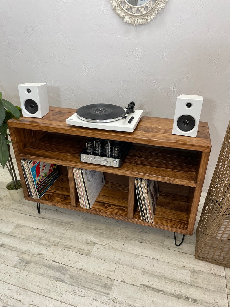 May include: A dark brown wooden record player stand with three shelves for storing vinyl records. The stand has black hairpin legs and features a white turntable and two white speakers.