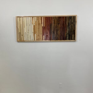 May include: A rectangular wooden wall art piece with a frame. The wood is arranged in vertical strips of different colors, ranging from light beige to dark brown.
