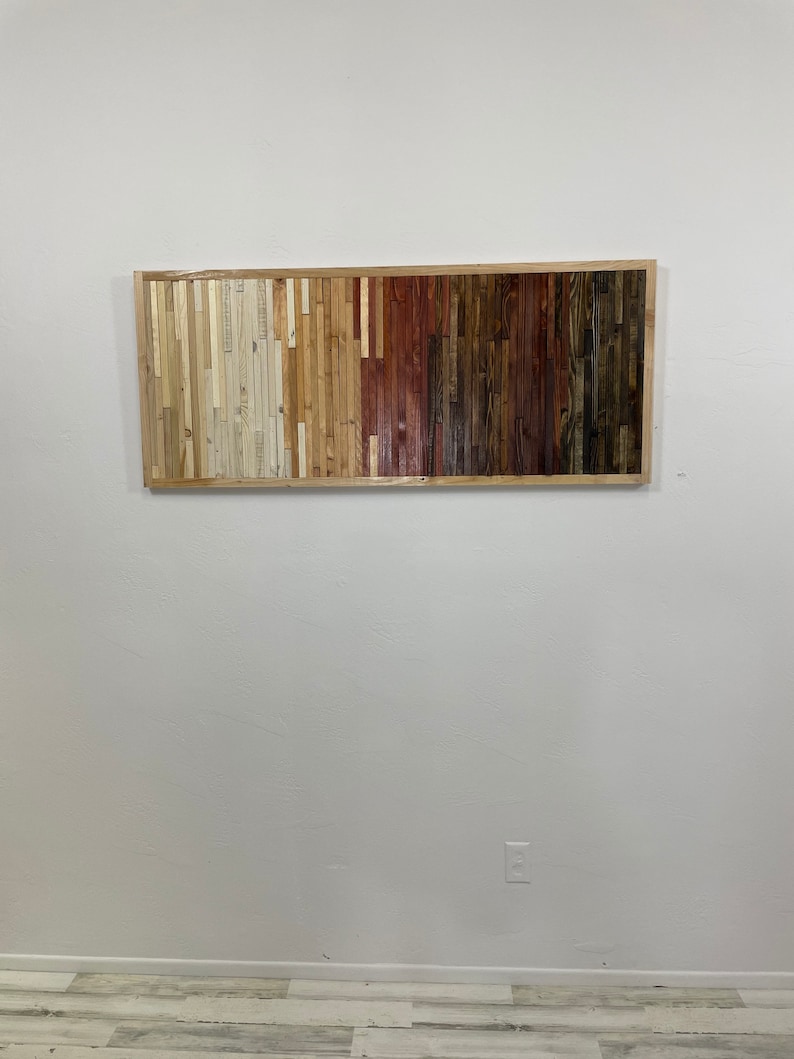 May include: A rectangular wooden wall art piece with a frame. The wood is arranged in vertical strips of different colors, ranging from light beige to dark brown.
