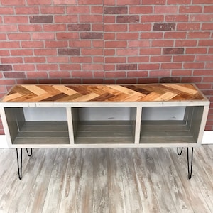 May include: A gray wooden shelf unit with three compartments and a herringbone patterned top. The shelf unit has black hairpin legs and is sitting on a light wood floor.