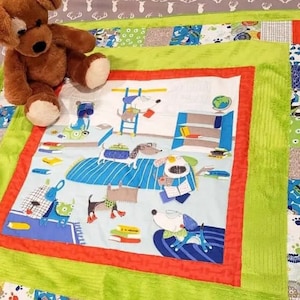 May include: A colorful baby quilt featuring a playful dog-themed design. The quilt has a central panel with cartoon dogs reading books, surrounded by a green border and patchwork squares. A brown teddy bear sits on top of the quilt.