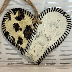 May include: A heart-shaped ornament made from two pieces of leather. One side is white with a distressed finish and the other side has a brown and white leopard print. Both sides have black stitching around the edges.