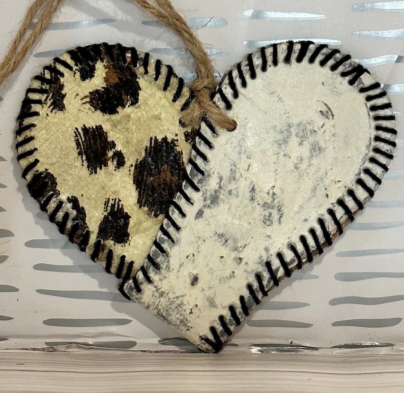 May include: A heart-shaped ornament made from two pieces of leather. One side is white with a distressed finish and the other side has a brown and white leopard print. Both sides have black stitching around the edges.