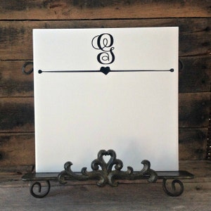May include: White ceramic tile with a black cursive letter "G" and a small heart above a black line. The tile is on a black metal stand with a decorative scroll design.