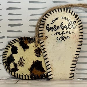 May include: White heart-shaped ornament with a baseball stitch design. The ornament is split in half, one side has a brown and white leopard print and the other side has the text "Livin' that baseball mom life".