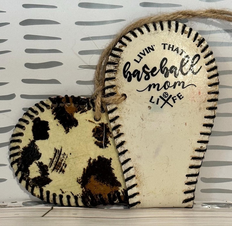 May include: White heart-shaped ornament with a baseball stitch design. The ornament is split in half, one side has a brown and white leopard print and the other side has the text "Livin' that baseball mom life".