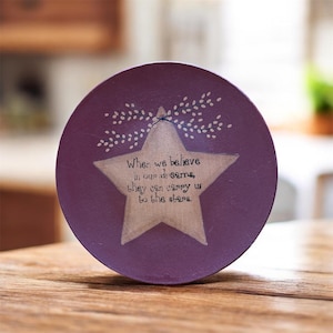 Może przedstawiać: Okrągły, fioletowy drewniany talerz z namalowaną gwiazdą i napisem "When we believe in our dreams, they can carry us to the stars." Gwiazda jest jasno-beżowa, a talerz ma rustykalny, ręcznie robiony wygląd.