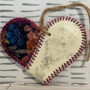 May include: A heart-shaped ornament with a white background and a pink stitched edge. One side of the heart is covered in a colorful floral fabric.