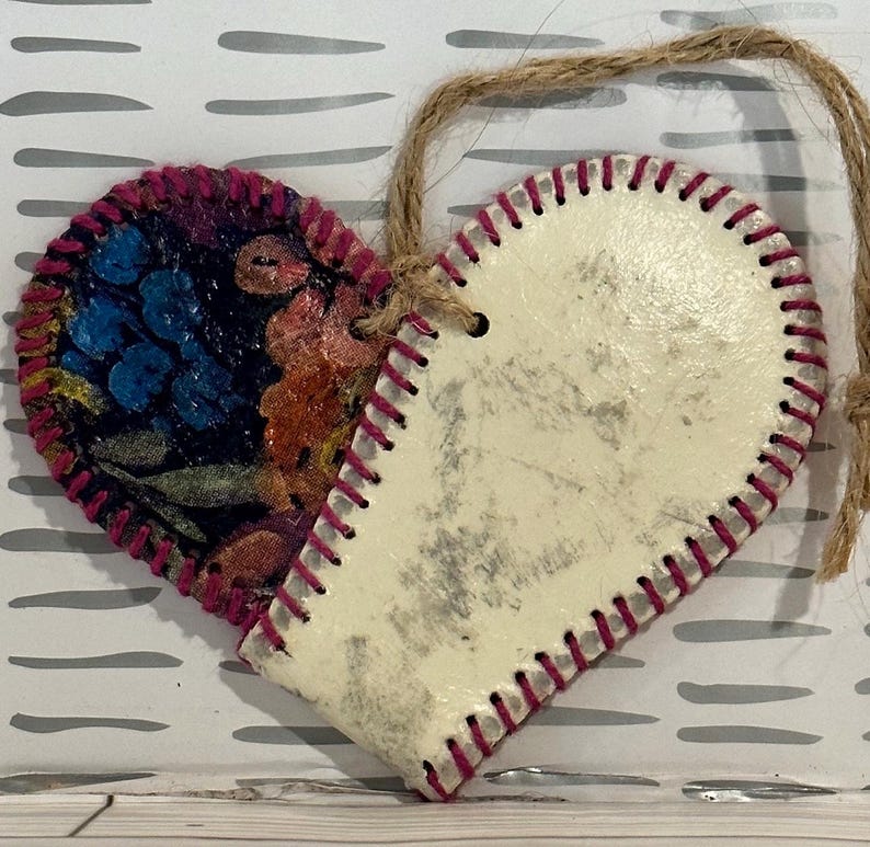 May include: A heart-shaped ornament with a white background and a pink stitched edge. One side of the heart is covered in a colorful floral fabric.