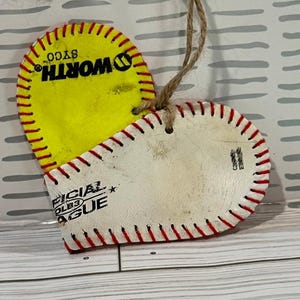 May include: A heart-shaped ornament made from two baseballs. The front is a white baseball with red stitching and the text "OFFICIAL COLEB3 LEAGUE". The back is a yellow baseball with red stitching and the text "WORTH SYCO".
