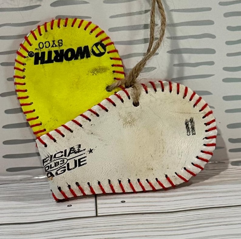 May include: A heart-shaped ornament made from two baseballs. The front is a white baseball with red stitching and the text "OFFICIAL COLEB3 LEAGUE". The back is a yellow baseball with red stitching and the text "WORTH SYCO".